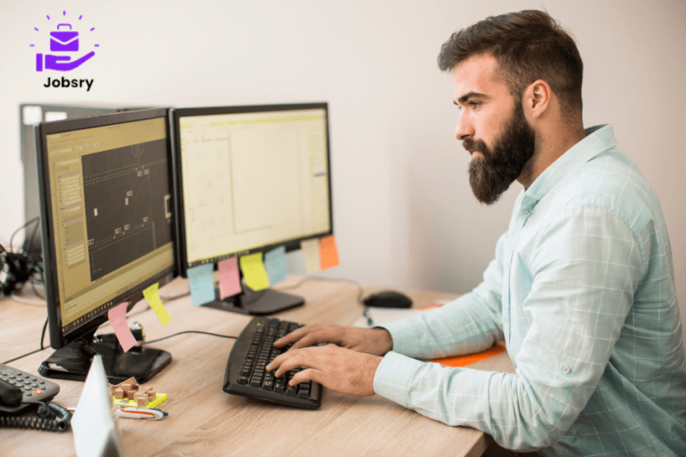 A bearded man sits at a desk with two computer screens, working as a computer operator while writing a job application letter.